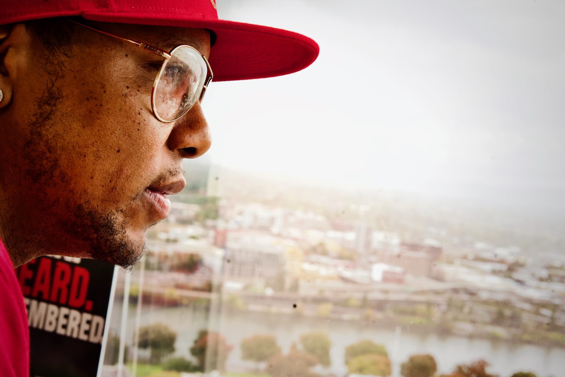 Close-up side profile of a man wearing a red hat and glasses looking out over a city skyline through a window, symbolizing focus and reflection.